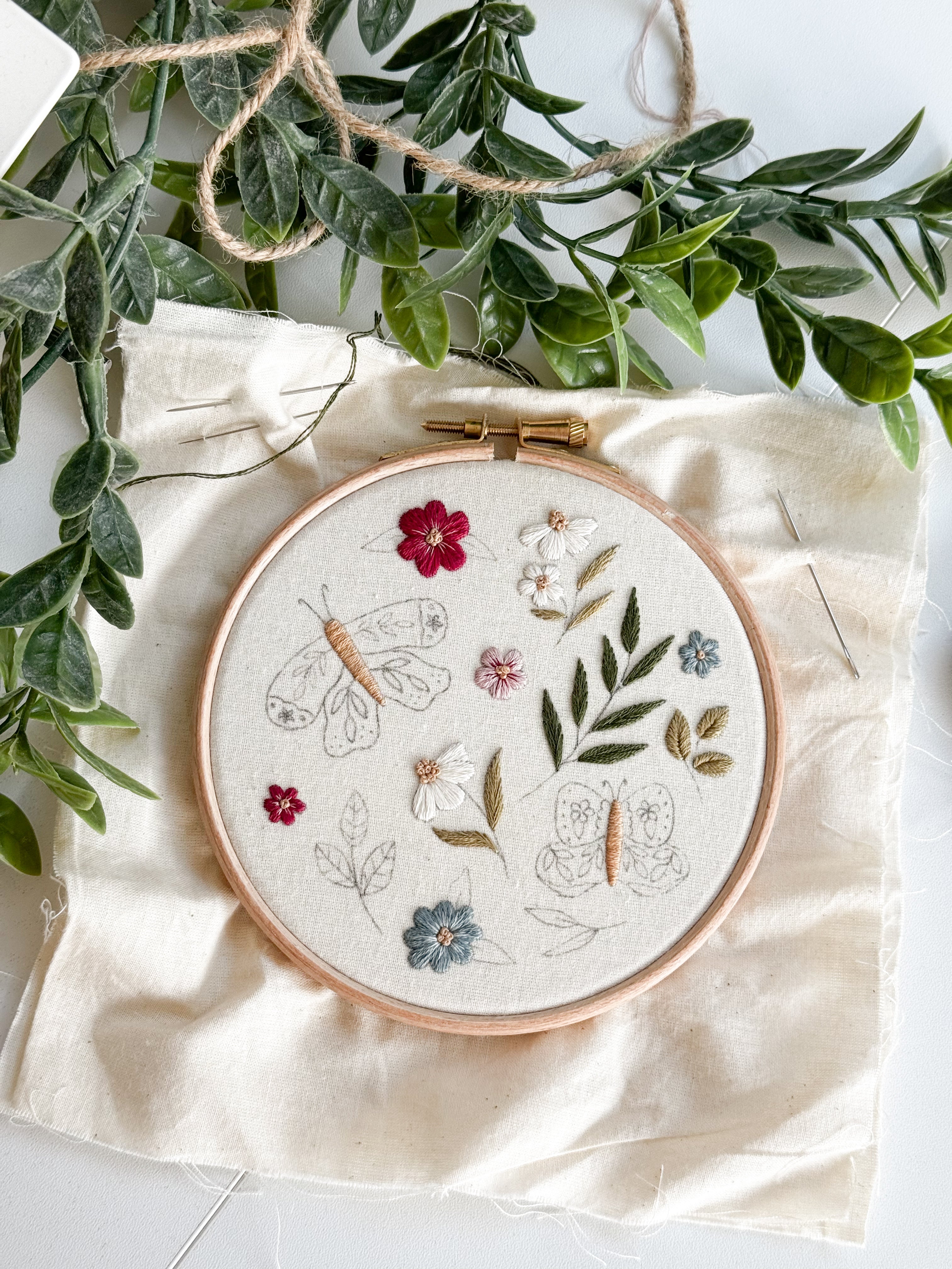 Embroidery hoop with floral and butterfly design on a white fabric background with green leaves.
