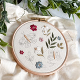 Embroidery hoop with floral and butterfly design on a white fabric background with green leaves.