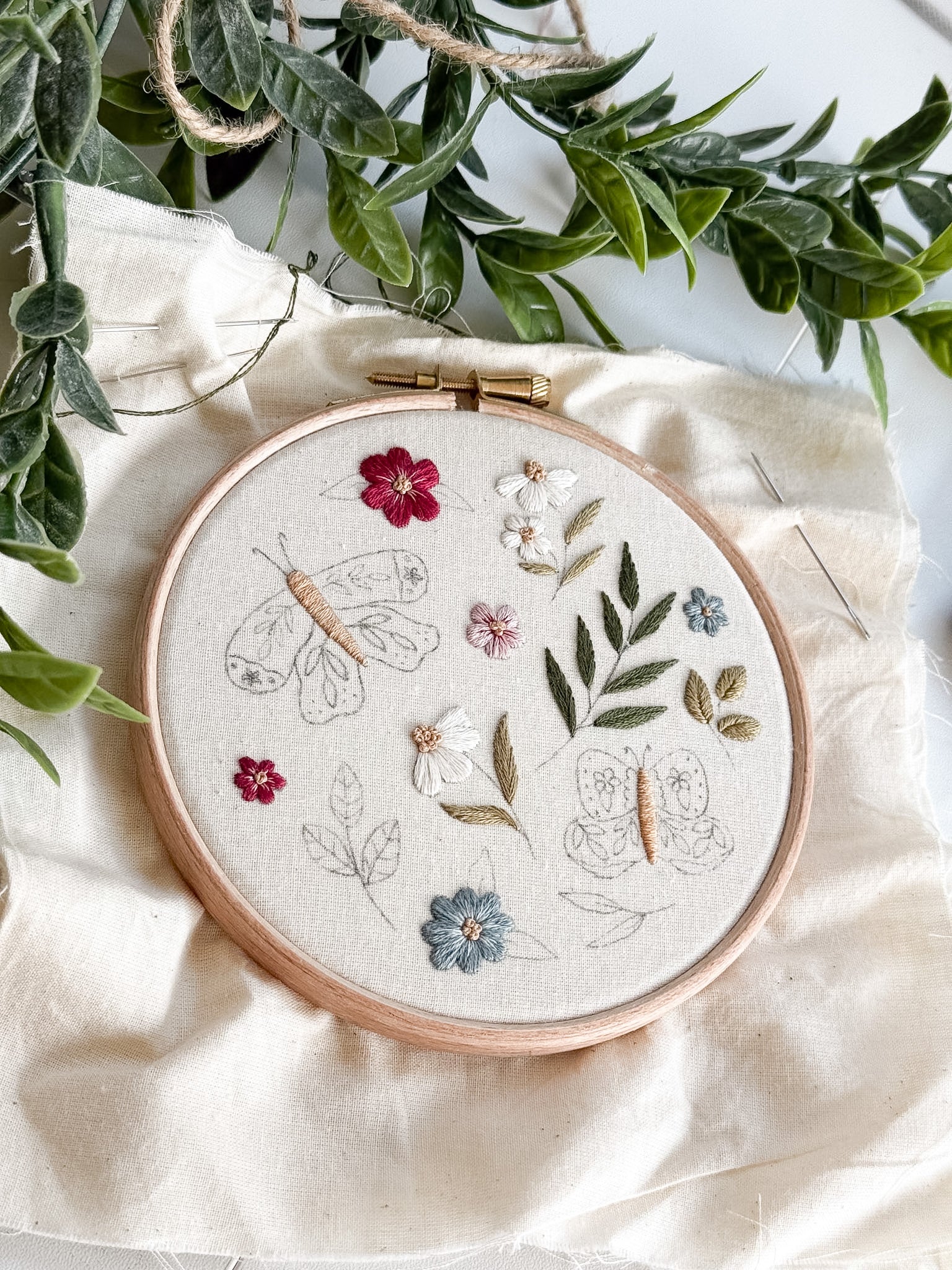 Embroidery hoop with floral and butterfly design on a white fabric background with green leaves.