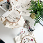 Embroidery hoops with floral designs on a table with a plant and sewing kit.