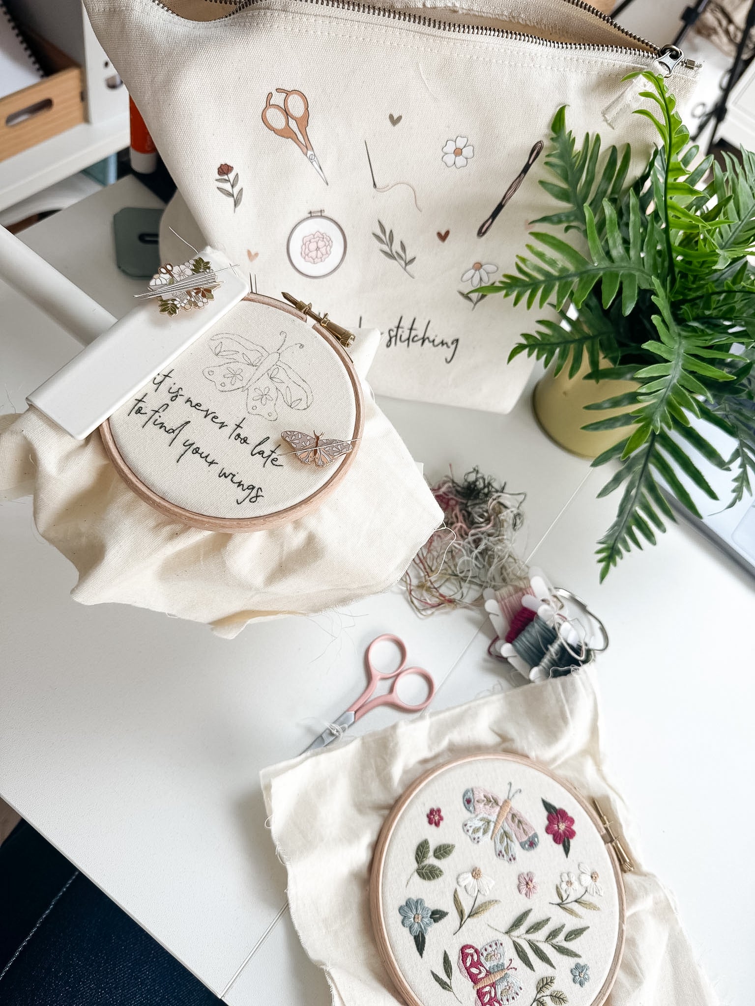 Embroidery hoops with floral designs on a table with a plant and sewing kit.