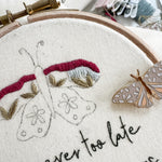 Embroidery hoop with embroidered butterflies and text 'It is never too late to change your wings'. A butterfly needle minder sits on the hoop