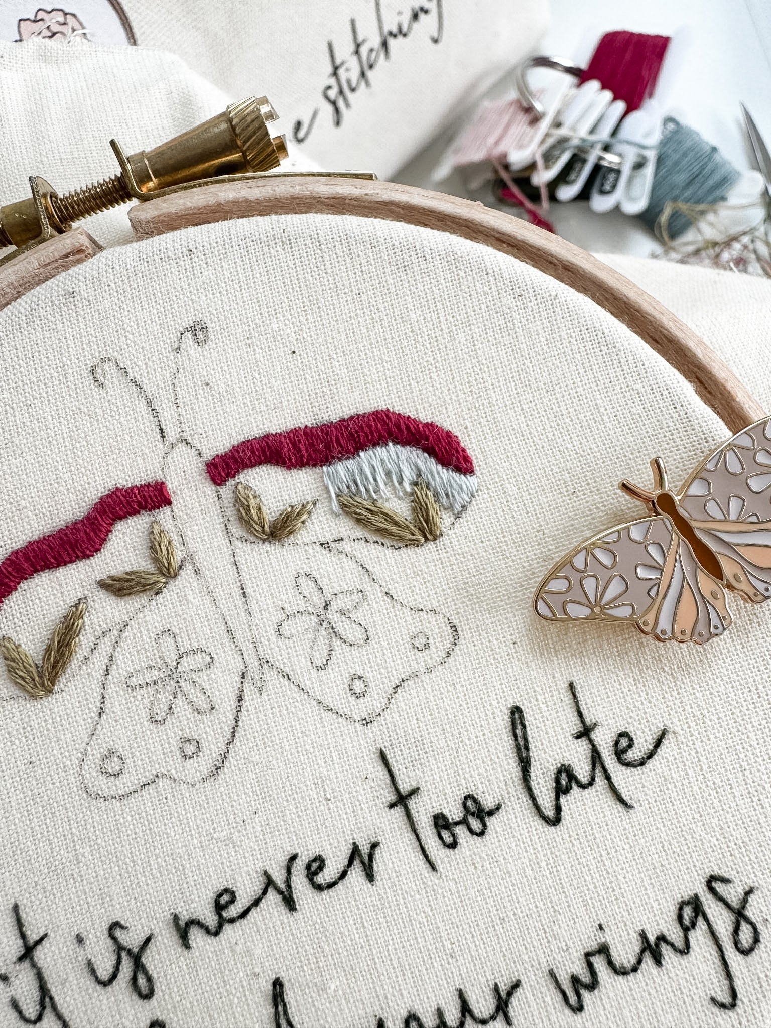 Embroidery hoop with embroidered butterflies and text 'It is never too late to change your wings'. A butterfly needle minder sits on the hoop