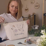 Woman holding a project bag with embroidery designs on a table with crafting supplies.
