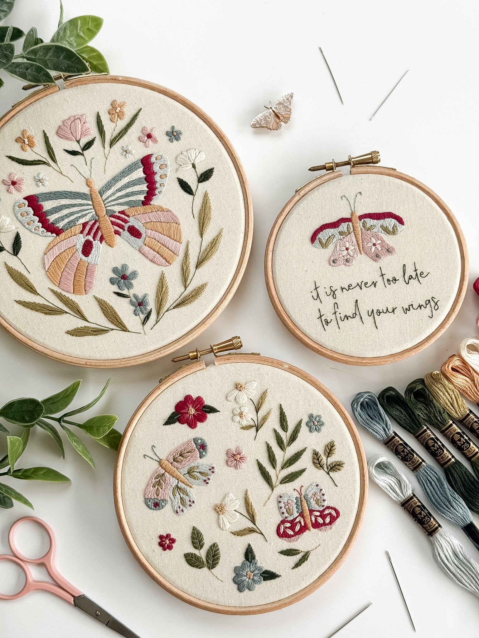 Three embroidered hoops with butterfly patterns on a white surface with embroidery threads and scissors.
