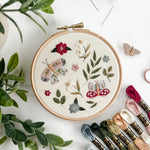 Embroidery hoop with floral and butterfly design on a white surface with embroidery threads and plants.