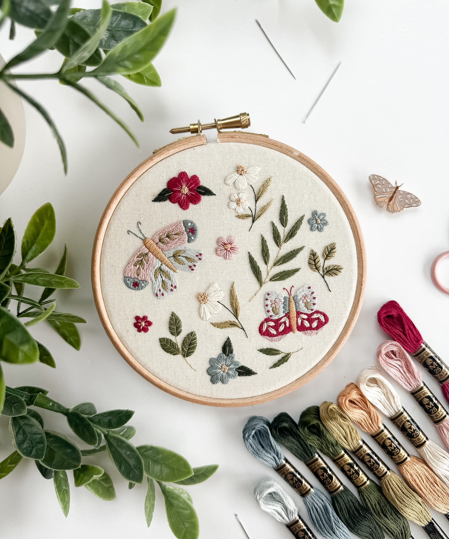 Embroidery hoop with floral and butterfly design on a white surface with embroidery threads and plants.