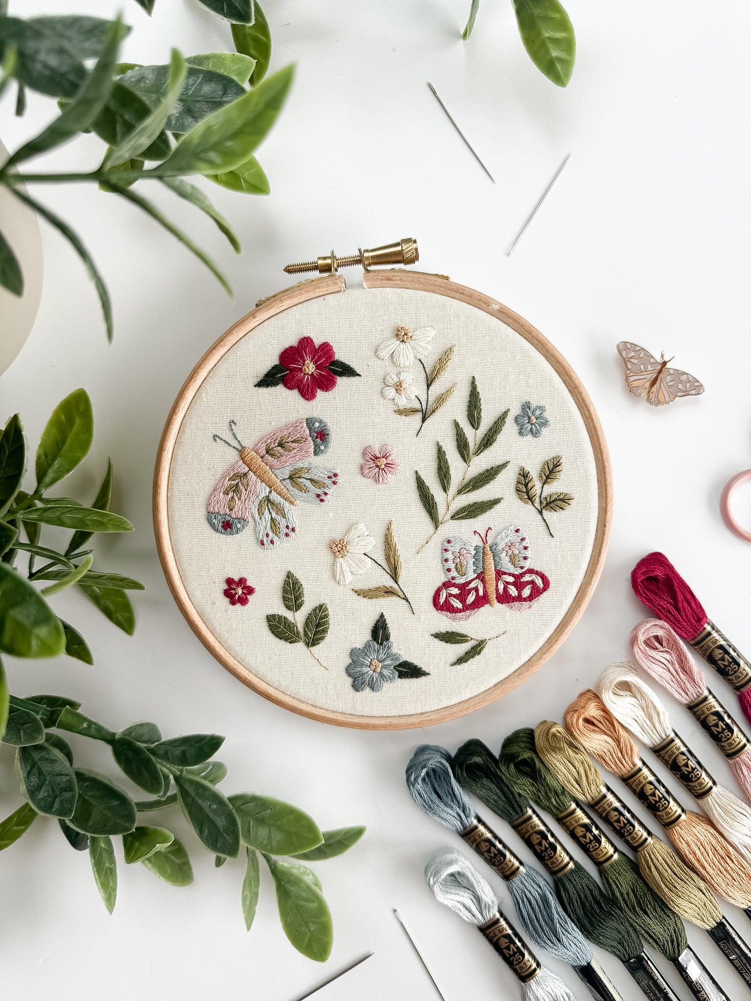 Embroidery hoop with floral and butterfly design on a white surface with embroidery threads and plants.