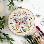 Embroidery hoop with butterfly design surrounded by embroidery threads and green leaves on a white surface