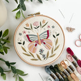 Embroidery hoop with butterfly design surrounded by embroidery threads and green leaves on a white surface