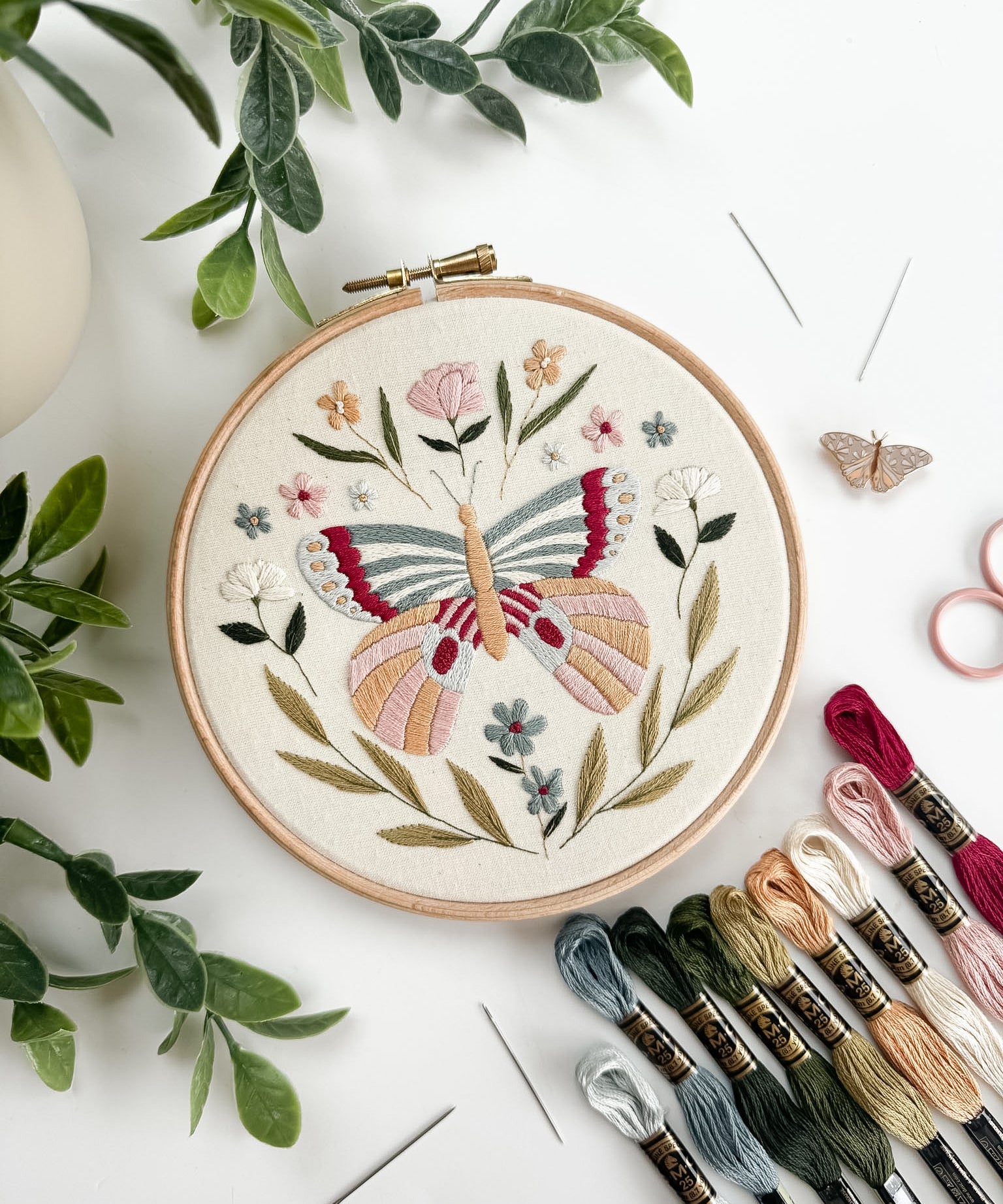 Embroidery hoop with butterfly design surrounded by embroidery threads and green leaves on a white surface