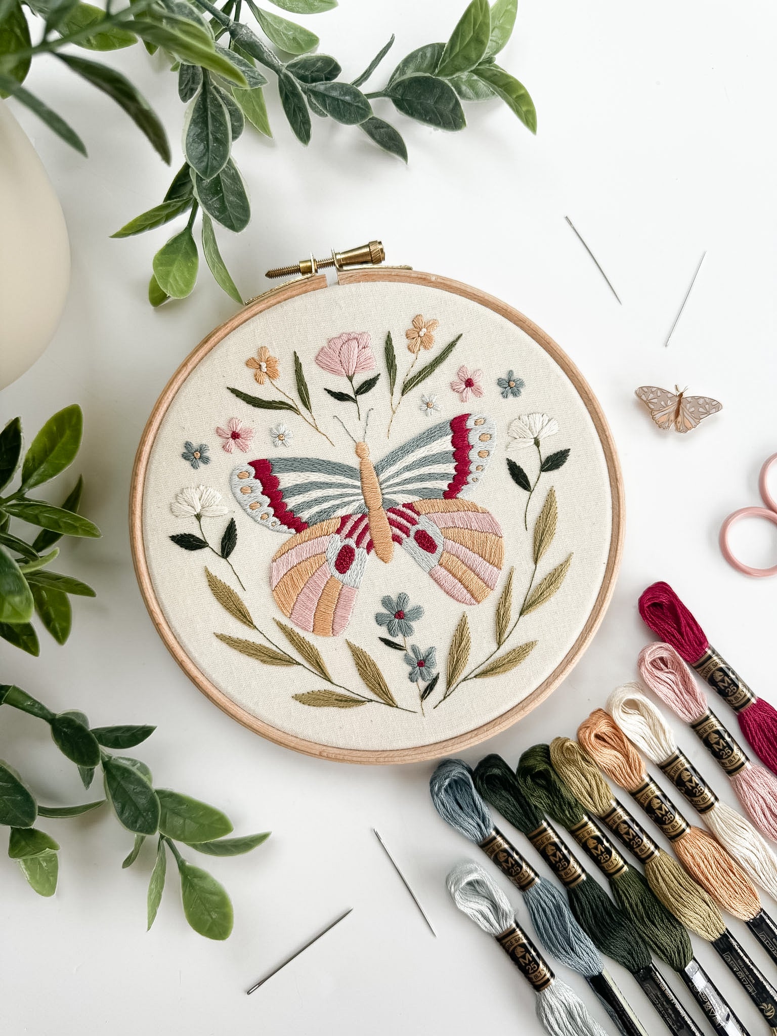 Embroidery hoop with butterfly design surrounded by embroidery threads and green leaves on a white surface