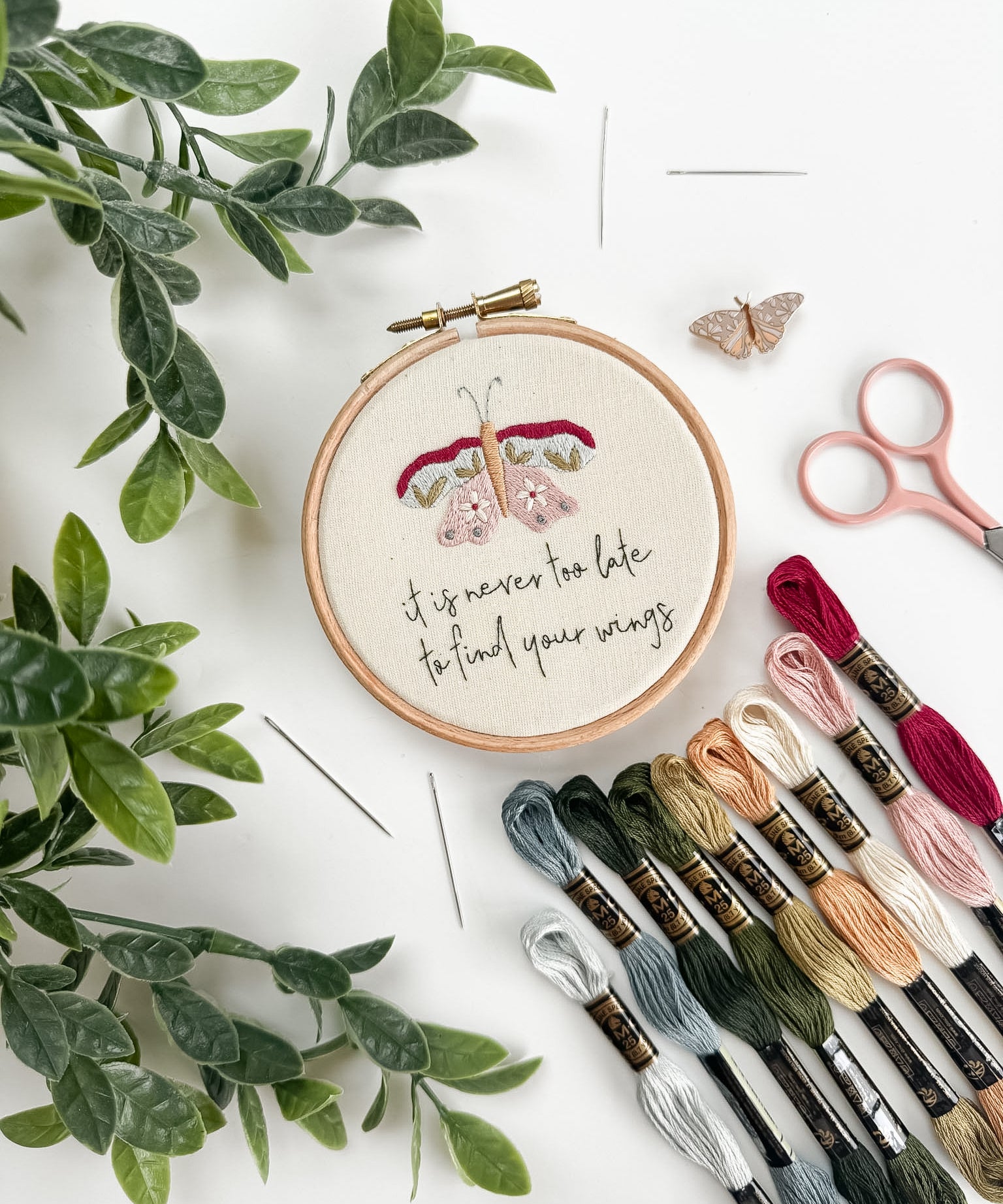 Embroidery hoop with floral design and motivational quote, surrounded by embroidery threads and scissors on a white background.