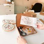 A woman opening a butterfly embroidery kit. She is holding the pattern booklet while 3 hoops are embroidered with butterflies on the table