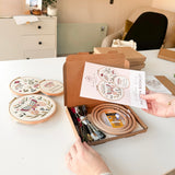 A woman opening a butterfly embroidery kit. She is holding the pattern booklet while 3 hoops are embroidered with butterflies on the table