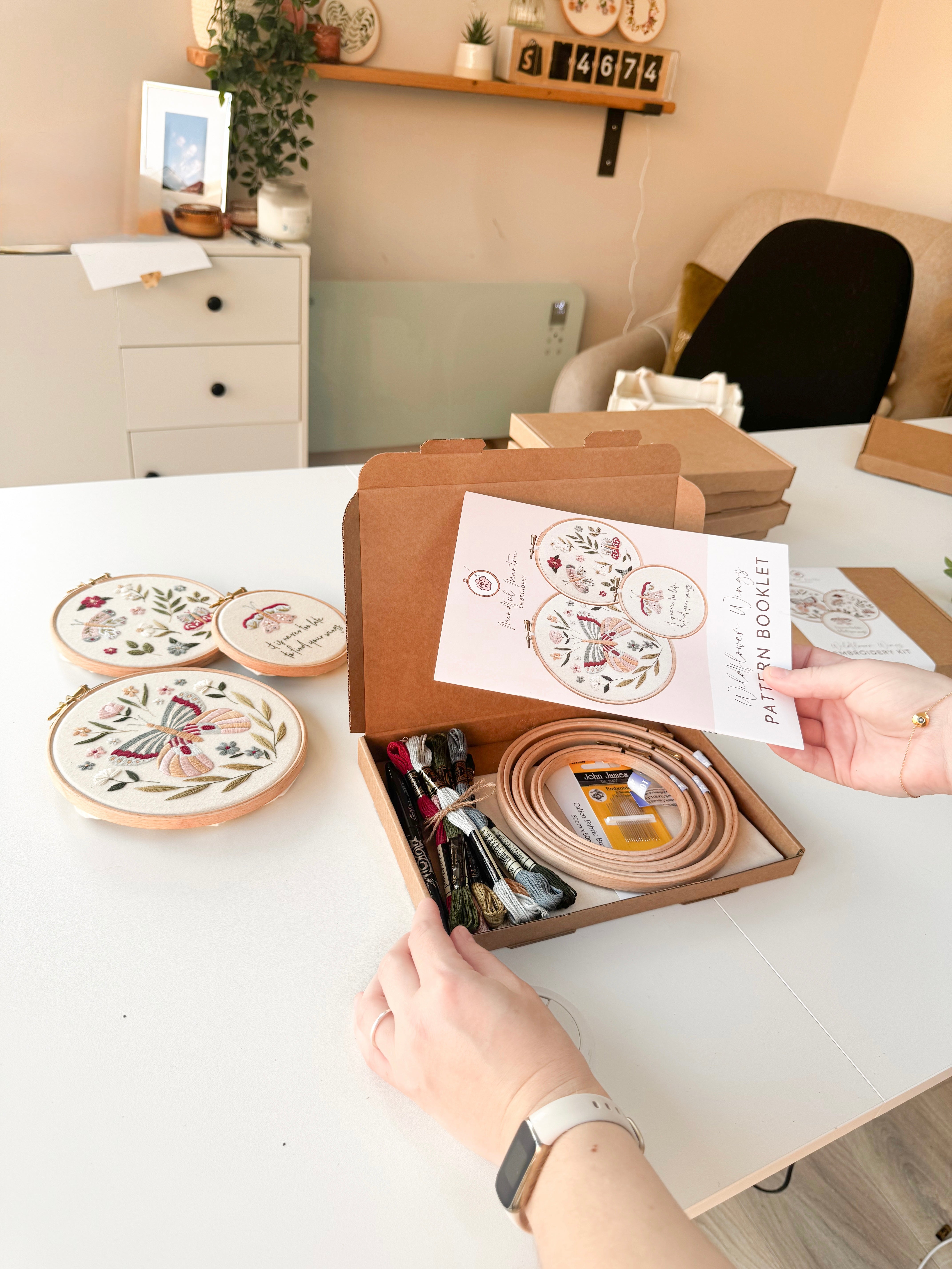 A woman opening a butterfly embroidery kit. She is holding the pattern booklet while 3 hoops are embroidered with butterflies on the table