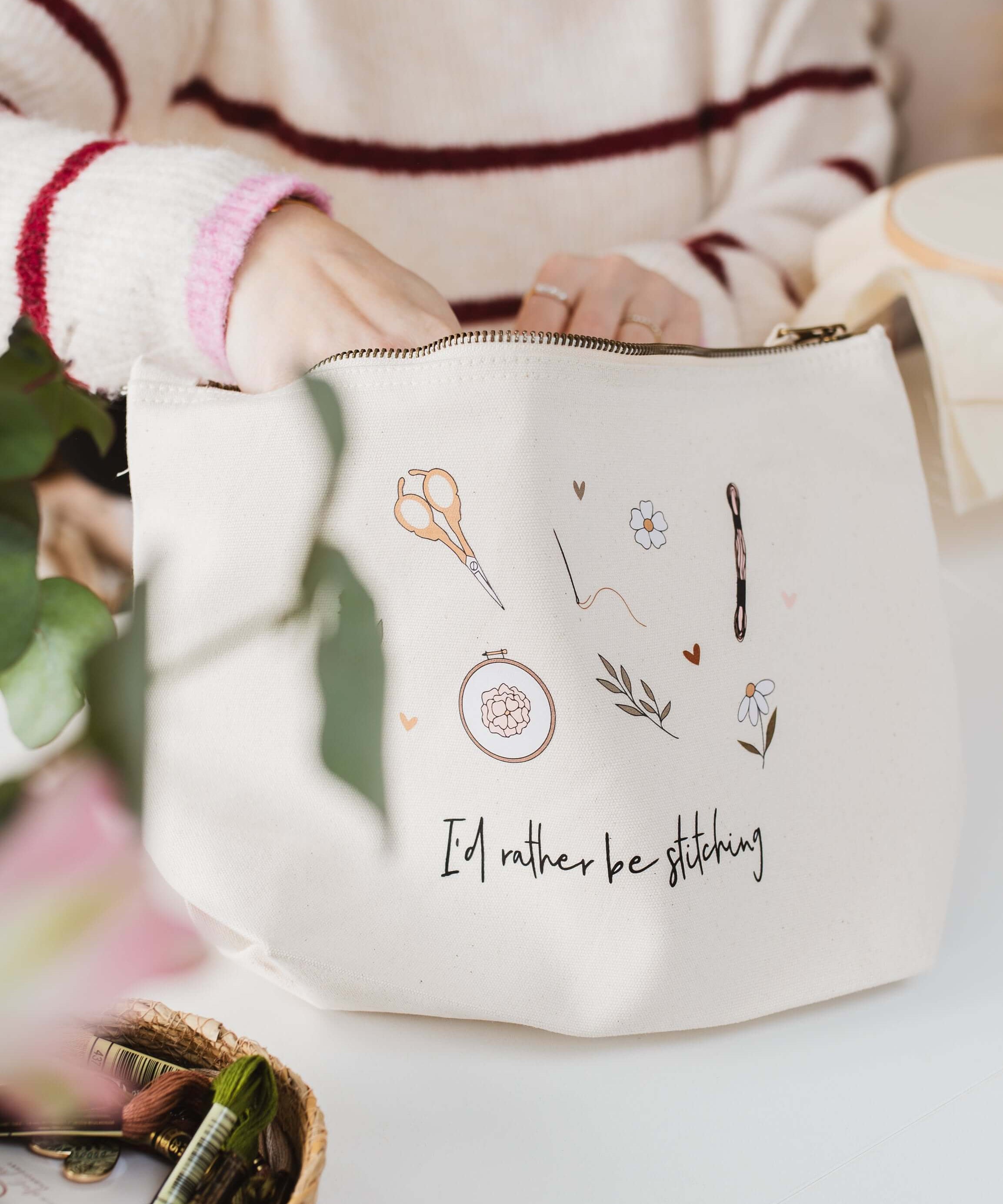 Person holding a white project bag with embroidery designs and 'I'd rather be stitching' text on a light background.