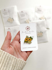 Magnetic Needle Minder featuring sunflowers, held in hand with packaging in background, perfect for needlework projects.